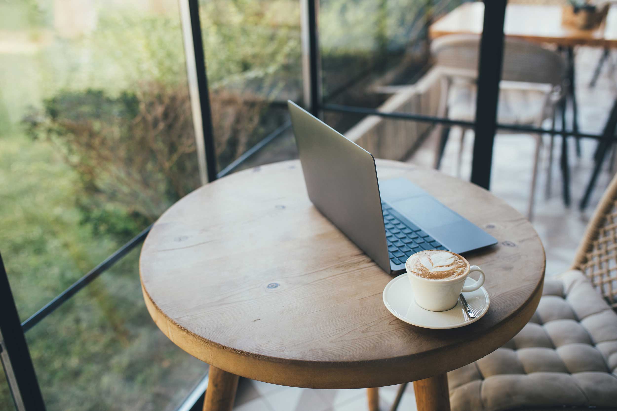 Schöner runder Holztisch mit passendem Stuhl am Fenster mit Laptop und Cappuccino.